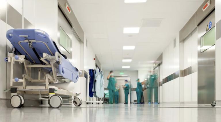 Bright hospital corridor with a wheeled patient bed in the foreground and medical staff in scrubs gathered near the end.
