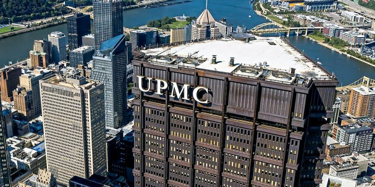 Aerial view of a downtown skyline with a tall brick building labeled UPMC beside a river.