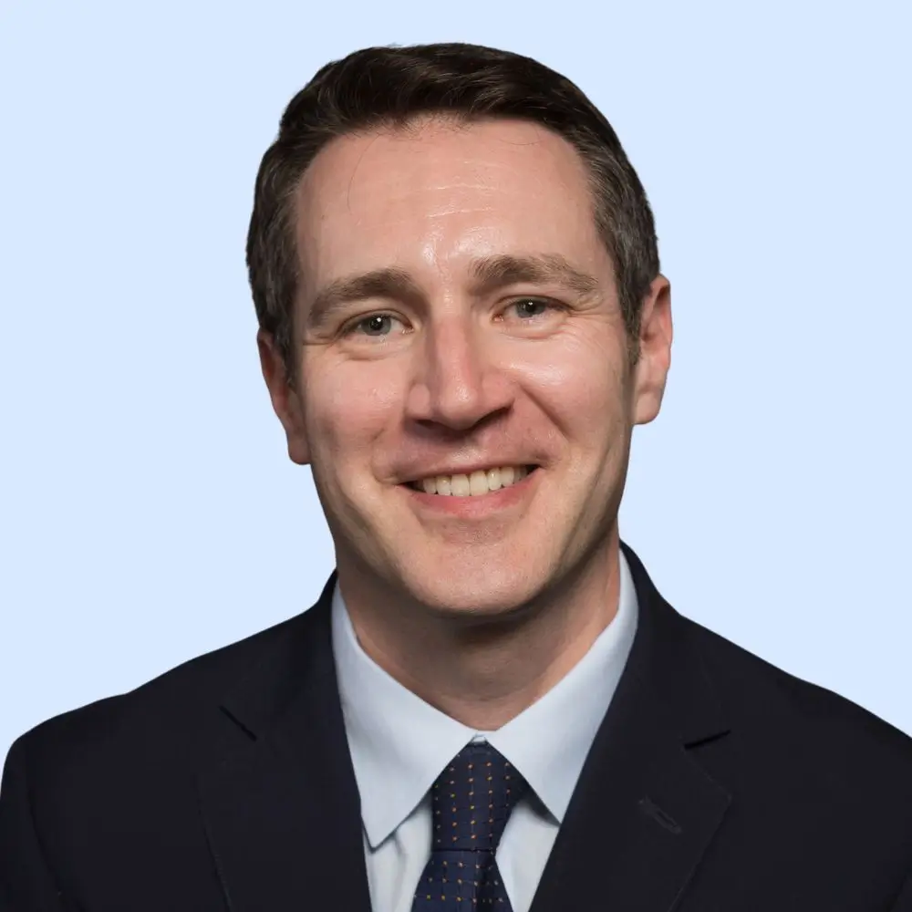 Smiling man in a dark suit and tie against a light blue background.